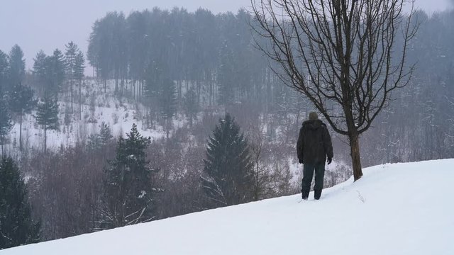 Man standing next to tree and watching snow falling on the forest - (4K)