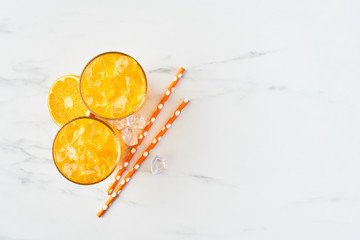 Fresh orange juice in tall glass with orange, ice and straws on white marble background. Summer drink. Top view. Copy space for text.