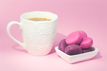 Several sweet tasty pink macarons in white bowl and white coffee mug on pink background with copy space closeup