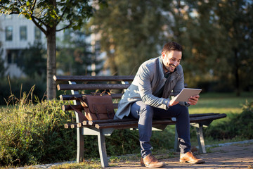 Positive Businessman at the park with tablet sitting on a bench