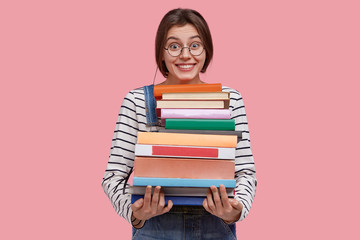 Delighted young woman carries pile of textbooks, smiles broadly, learns useful information from encyclopedia, has dark hair, dressed in casual outfit, poses aginst pink background. Education concept