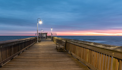 Fototapeta premium Sunrise from the Sandbridge Fishing Pier on Little Island Park in Virginia Beach. The wood of the pier is lit by early morning sun rising in the clouds, creating pink and purple light.