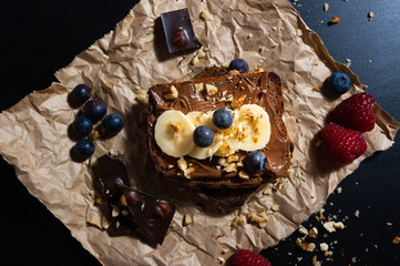  Delicious isolated chocolate bread toast with chocolate spread, banana, bluberry, rasberry and nuts toppings on rustic paper with black background