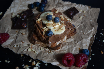  Delicious isolated chocolate bread toast with chocolate spread, banana, bluberry, rasberry and nuts toppings on rustic paper with black background