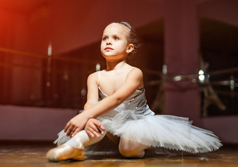Little ballerina in white tutu sitting on floor and resting in dance studio. Pretty small girl in ballet suit sits on the wooden floor and dreaming © Vadim