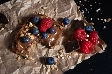 Bite of delicious isolated chocolate bread toast with chocolate spread, fresh banana, bluberry, rasberry and nuts toppings on rustic paper with black background