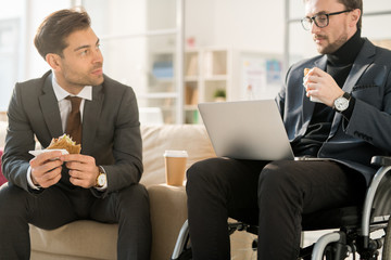 Young businessman in wheelchair using laptop while eating sandwich together with his colleague at office