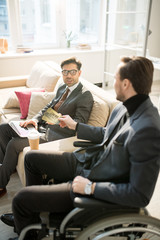 Fototapeta premium Businessman sitting in wheelchair and giving two tickets in cinema to his colleague while he sitting on sofa and using laptop