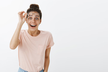 Excited and surprised pleased young beautiful woman with tan and messy hairbun taking off glasses as meeting friend and feeling happy smiling broadly with dreamy positive expression over gray wall