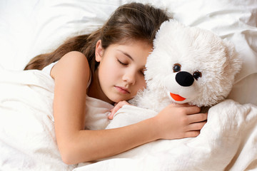 Cute little girl with long hair sleeping with teddy bear in bed. Adorable small child lies on warm comfortable bed having good dreams embracing her favourite soft toy at night