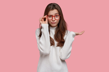 Photo of displeased serious young lady looks scrupulously through spectacles, keeps palm raised, wears oversized jumper, isolated over pink background. Hmm, look there. Advertisement concept