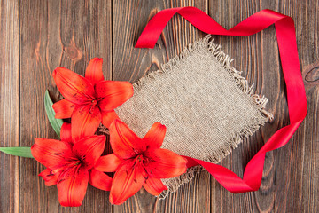 Red lilies with red ribbon and white paper blank on dark wooden background. Holidays card. 
