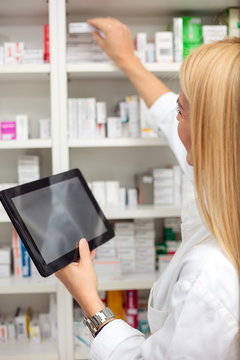 Smiling young female pharmacist holding a tablet and reaching for medications on a shelf, working in a pharmacy