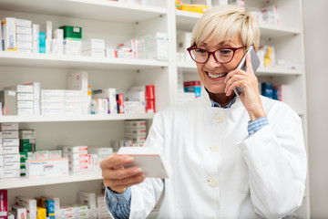 Happy senior female pharmacist checking medication details on a box and talking on a smart phone....