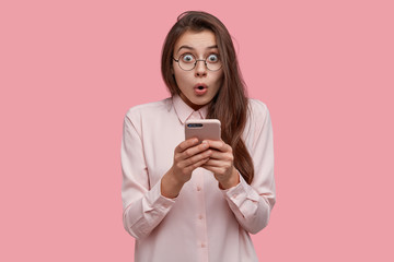 Waist up shot of shocked Caucasian lady holds mobile phone, surprised to read spam, made mistake while typing message, recieves high phone bills, dressed in elegant shirt, isolated on pink wall - obrazy, fototapety, plakaty