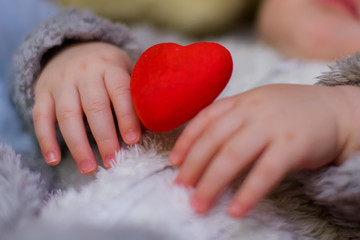 The small arms of a newborn child hold a red heart. The child of the parents was kidnapped. Kid on Valentine's Day. Postcard to Mother's Day.