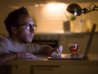 Concentrated Caucasian man designer with eyeglasses in beige sweater and cup of tea working / typing on laptop computer at night home. Freelance, overwork, deadline, remote work concept.  