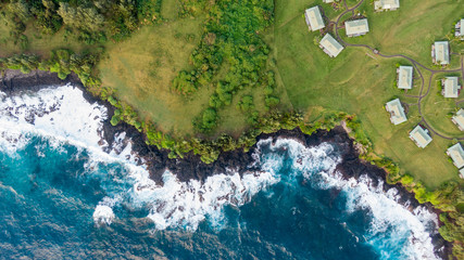 Stunning aerial view of the rough volcanic coastline in the town of Hana on the eastern side of the island of Maui, Hawaii. Beautiful waves, volcanic rocks, grassland, trees and bungalow houses.
