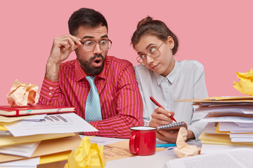 Teamwork concept. Young coworkers in big glasses brainstrom together, study documents for future business deal, write down notes in spiral notepad, drink hot beverage, isolated on pink background