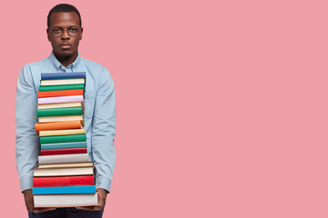 Studio shot of dark skinned serious confident college student carries many books in pile, wears glasses and shirt, returns from library, chooses literature for writing course paper, isolated on pink