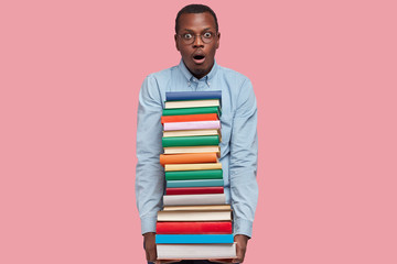 Shot of surprised African American man carries pile of colourful books, scared of having deadline, dressed in formal wear, keeps jaw dropped, isolated over pink background, analyzes literature