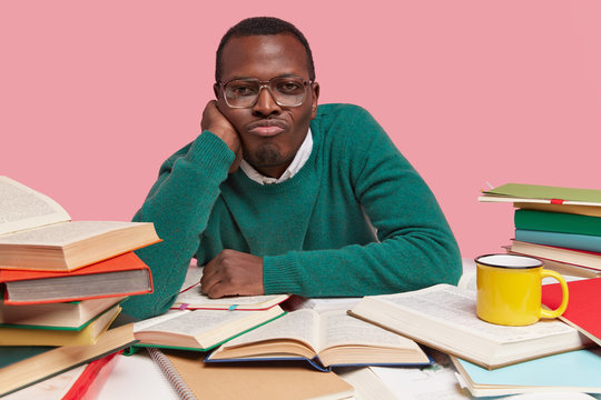 Photo of unhappy black young man keeps hand under chin, purses lips, wears optical glasses, feels lonely, reads books, dressed in casual clothes, poses over pink background. Gloomy ethnic reader