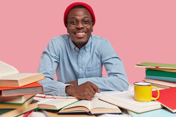 Cheerful black hipster guy in red headgear has toothy smile, wears shirt, cramms for exam, studies literature, uses book for getting knowledge, wears in elegant clothes, isolated over pink wall