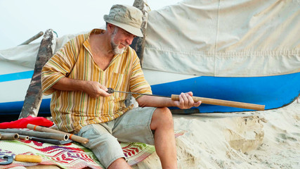 Senior man sits on the beach and making flute by hands.