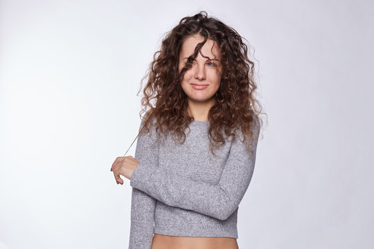 Youth and happiness concept. Close up of beautiful Caucasian lady in gray sweater, looking aside, playing with long curly gorgeous hair. Pretty woman with perfect natural appearance isolated indoors.