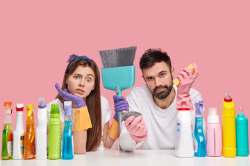 Displeased Caucasian woman and man look with displeasure, feels fatigue after spring cleaning in house, use broom, detergents and sponge for bringing apartment in order, isolated over pink wall
