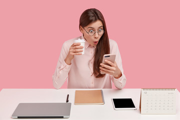 Photo of attractive young woman reads shocking news on mobile phone, watches video in social networks, drinks coffee from disposable cup, being office perfectionist, uses electronic gadgets.