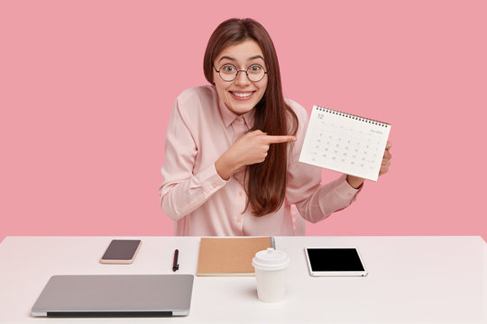 Positive young freelance worker points at calendar, demonstrates date of finishing project work, dressed in fashionable clothes, poses at desktop, isolated over pink background, enjoys hot drink