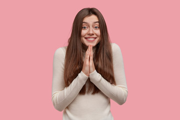 Pleased smiling lady has pleading expression, keeps palms pressed together, prays for everything...