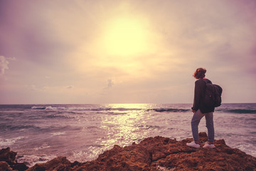 Fototapeta premium A girl with a backpack is standing with her back on the stormy seashore, an image with retro toning
