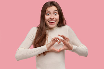Love and care concept. Beautiful woman smiles broadly, shows heart sign over chest, wears casual clothes, has happy exxpression, stands against pink background, expresses sympathy, feels uplifted