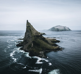 Abstract rock formations and an island covered with green grass and moss in rough sea from bird perspective on a moody day with rain and the sky as a blue background on the Faroe Islands in Europe