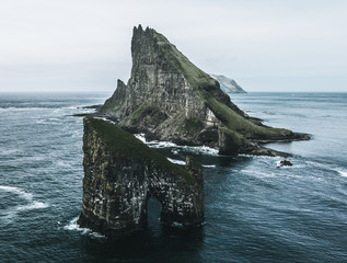 Abstract rock formations and an island covered with green grass and moss in rough sea from bird perspective on a moody day with rain and the sky as a blue background on the Faroe Islands in Europe