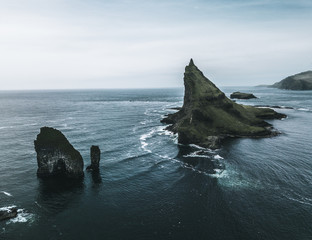 Abstract rock formations and an island covered with green grass and moss in rough sea from bird perspective on a moody day with rain and the sky as a blue background on the Faroe Islands in Europe
