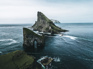 Abstract rock formations and an island covered with green grass and moss in rough sea from bird perspective on a moody day with rain and the sky as a blue background on the Faroe Islands in Europe