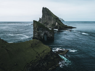 Abstract rock formations and an island covered with green grass and moss in rough sea from bird perspective on a moody day with rain and the sky as a blue background on the Faroe Islands in Europe