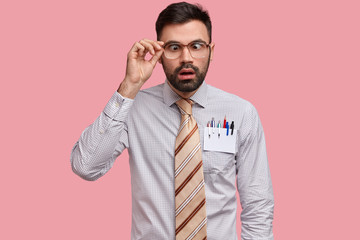 Surprised dark haired young man crosses eyes, keeps hand on rim of spectacles, dressed in formal clothes, keeps mouth opened, isolated over pink background. Funny male director makes grimace