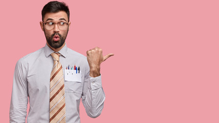Portrait of attractive bearded guy in formal clothes, looks in amazement aside, points at blank...