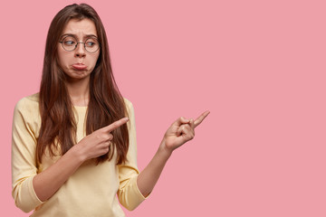 Depressed young woman purses lower lip, indicates at something on pink studio wall, has displeased facial expression, looks with dissatisfaction and frustration, doesnt like item. Look aside