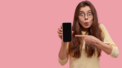 Astonished brunette young woman points at smart phone with black empty screen, surprised with many...