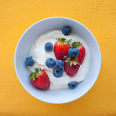Yoghurt with fresh strawberries and blueberries in bowl on yellow background, top view