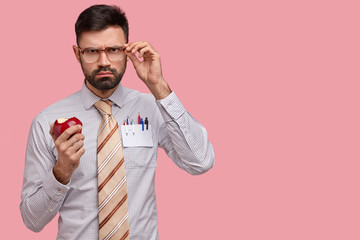 Serious gloomy male boss in formal clothes keeps hand on spectacles, bites delicious red apple, isolated over pink background with copy space for your advertisement or promotion, has snack after work