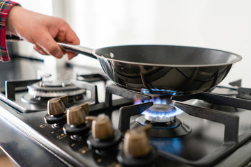 A man cooks in a frying pan, puts it on the stove. Modern gas burner and hob on a kitchen range. Dark black color and wooden Small kitchen in a modern apartment - obrazy, fototapety, plakaty