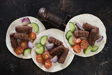 Fototapeta na wymiar Barbecued balkan cevapcici sausages with vegetables on tortilla wraps, flatlay on a dark brown stone background