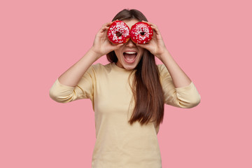 Studio shot of happy dark haired woman covers eyes with two red donuts, being in high spirit, wears yellow clothes, eats delicious junk food, has fun with friends. Female youngster holds doughnuts