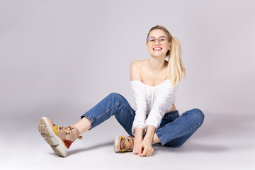 Studio shot of gorgeous young blonde woman with straight hair wearing off shoulder crop top sweater and high waisted denim shorts. Gray isolated background, copy space, close up. Ugly sneakers concept